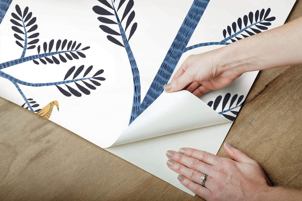 Hands peeling back Aviary Branch blue peel and stick wallpaper showing its botanical pattern.
