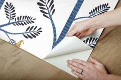 Hands peeling back Aviary Branch blue peel and stick wallpaper showing its botanical pattern.