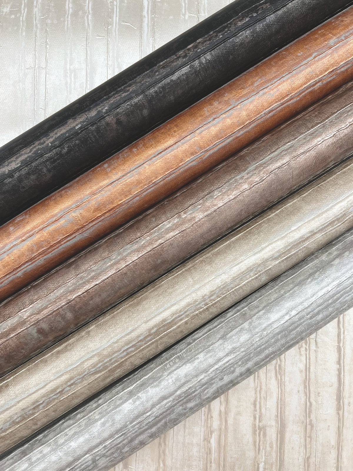 Five rolls of Ronald Redding Vintage Tin Wallpaper in black, copper, bronze, gold, and silver arranged diagonally against a patterned background.