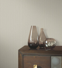 Three glass vases sit on a dark wooden cabinet against a backdrop of off-white Ronald Redding Vintage Tin Wallpaper featuring a subtle, vertical textured pattern.