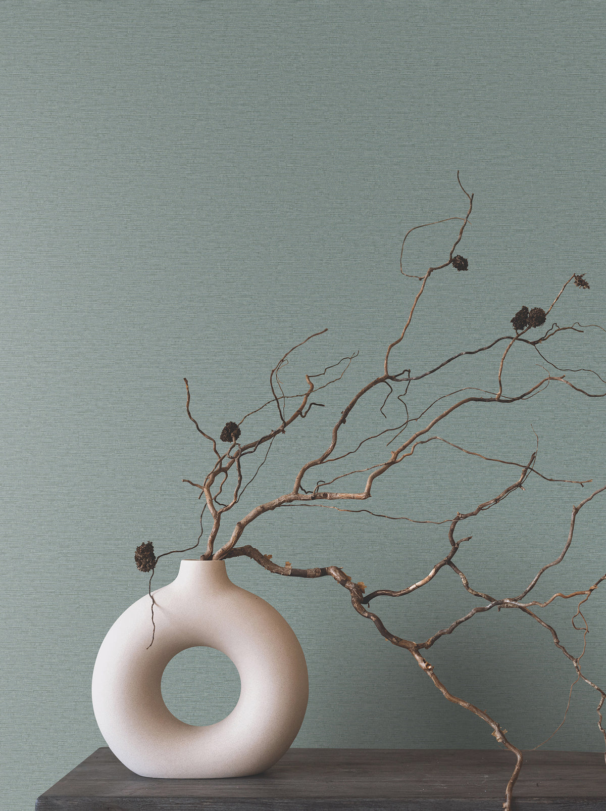 Matte, sage green Ronald Redding Cantilever Wallpaper with a subtle horizontal texture behind a white, ring-shaped vase holding twisted branches.