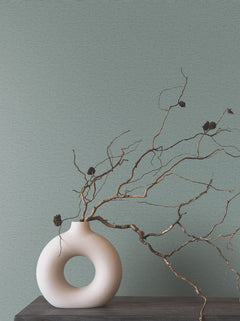 Matte, sage green Ronald Redding Cantilever Wallpaper with a subtle horizontal texture behind a white, ring-shaped vase holding twisted branches.
