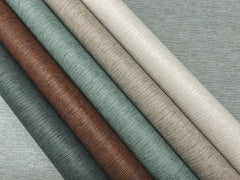 Five rolls of Ronald Redding Cantilever Wallpaper in shades of gray, brown, green, and beige laid diagonally across a gray, textured background.