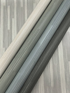 Three rolls of Ronald Redding Newel Wallpaper by York Wallcoverings, in shades of light beige, gray, and dark gray, are laid diagonally on a matching striped metallic background.