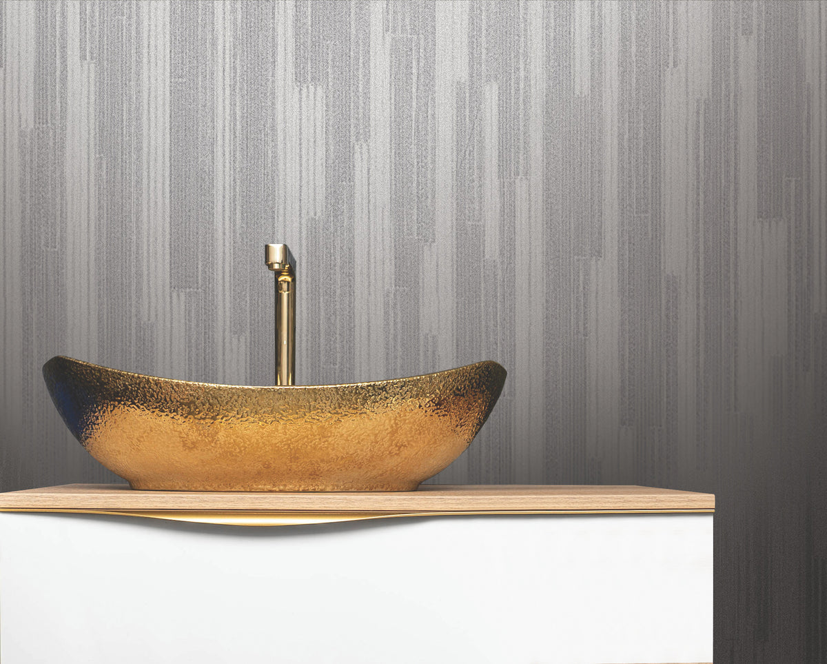 A gold textured vessel sink with a tall gold faucet sits on a white countertop against a wall adorned with Ronald Redding Newel Wallpaper by York Wallcoverings.