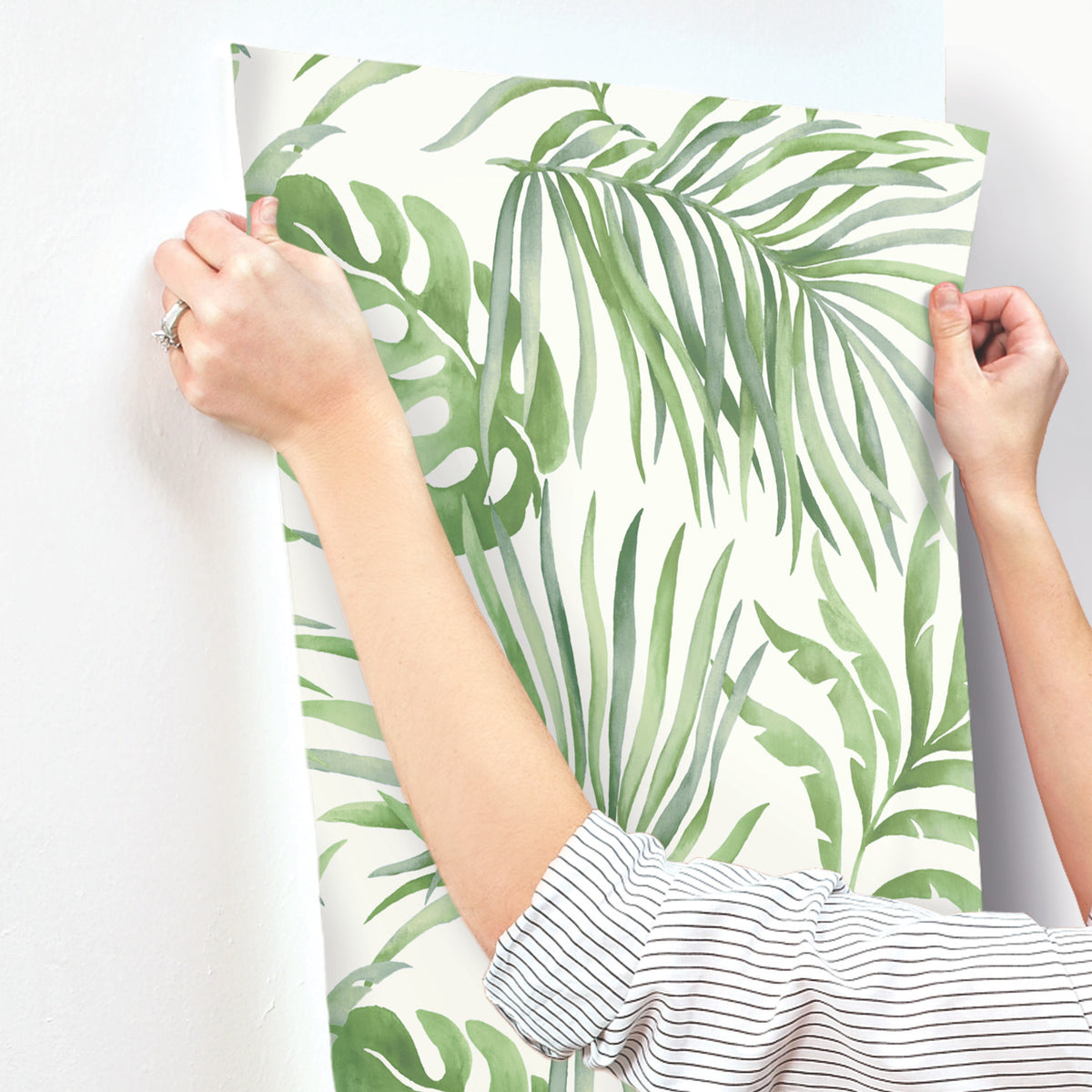 Hands applying Paradise Palm Wallpaper, featuring a green watercolor palm leaf pattern, to a white wall.