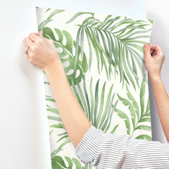 Hands applying Paradise Palm Wallpaper, featuring a green watercolor palm leaf pattern, to a white wall.
