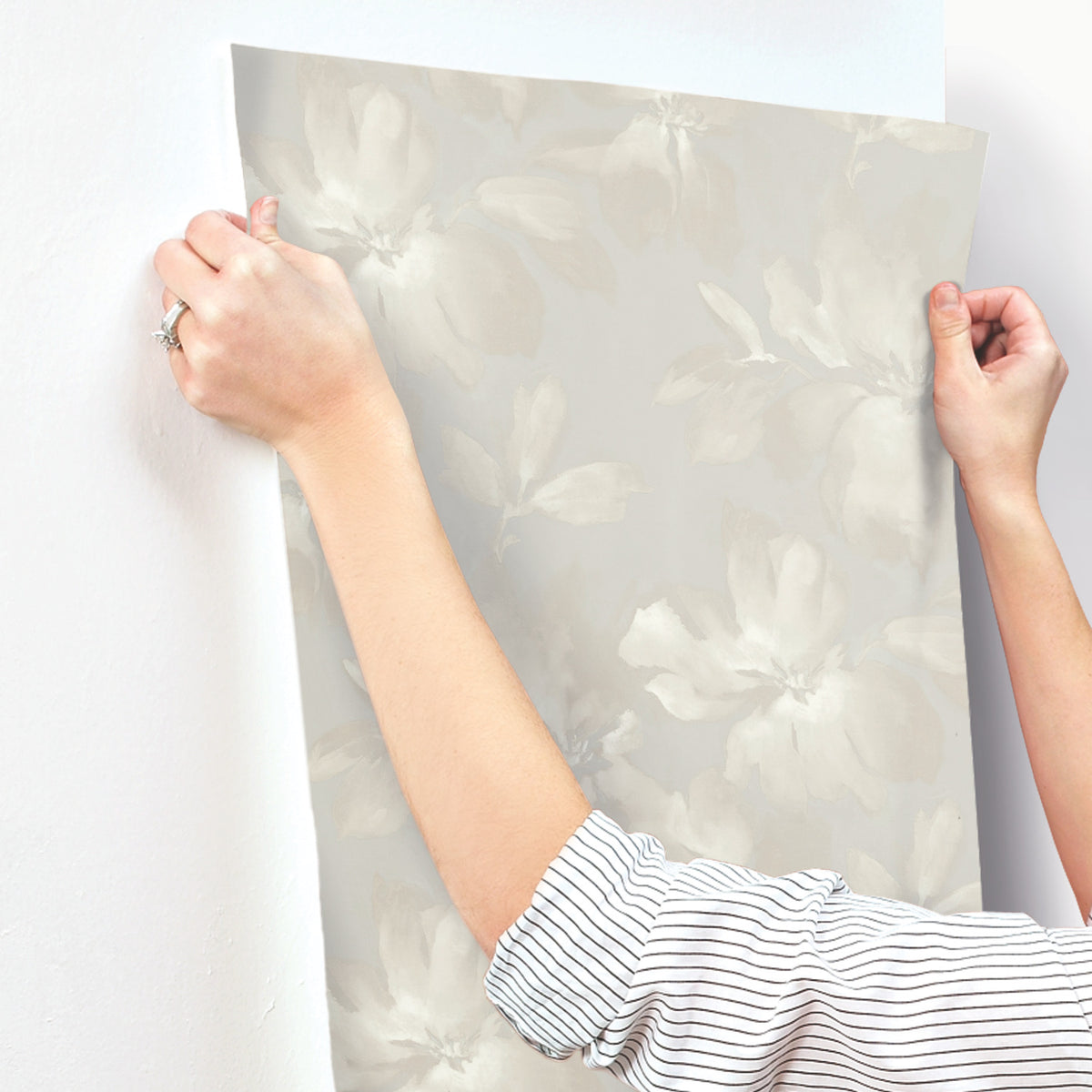 Hands applying a sheet of light gray Midnight Blooms Wallpaper featuring a large-scale floral pattern to a wall.