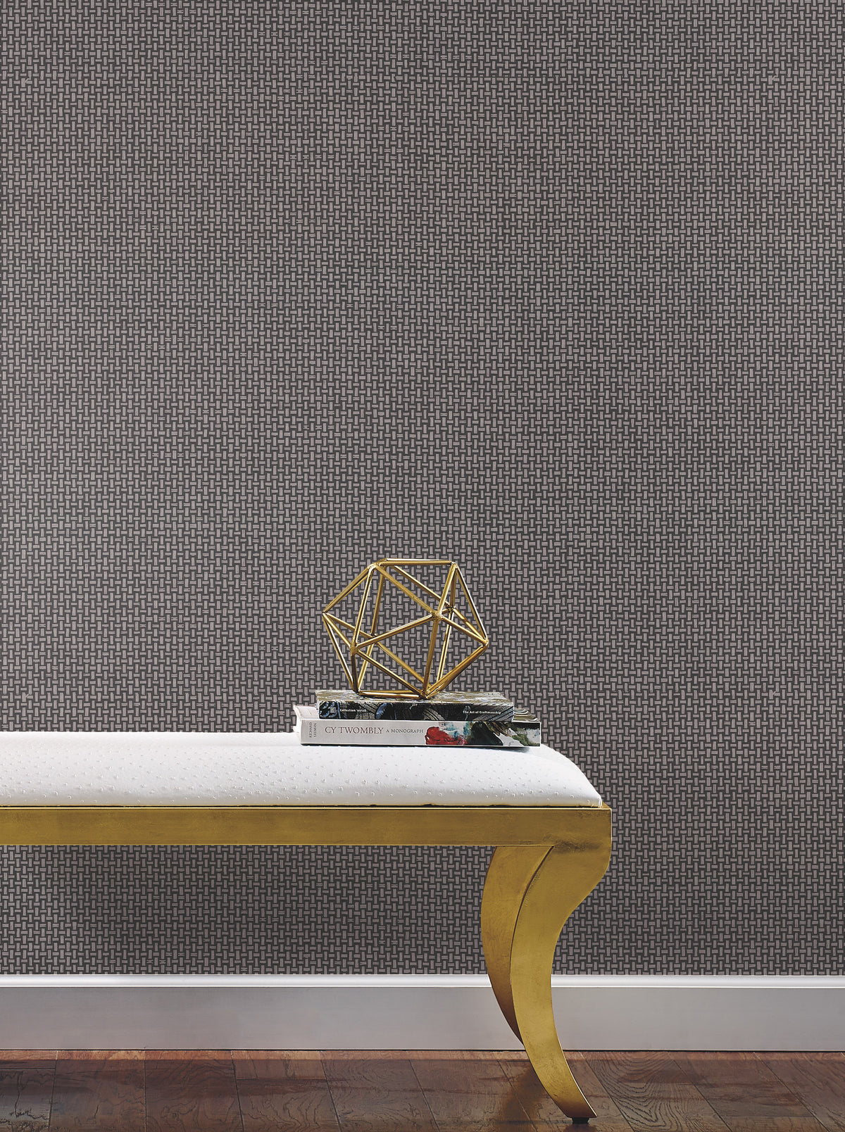 A gold-legged bench with a white cushion sits on a wooden floor. On the bench are two stacked books, a pair of glasses, and a gold geometric sculpture. The background features York Wallcoverings’ Cottage Basket Wallpaper in textured, patterned grey.