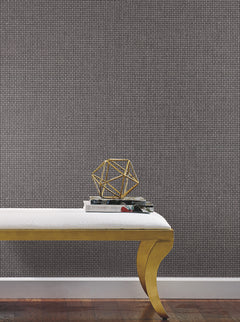 A gold-legged bench with a white cushion sits on a wooden floor. On the bench are two stacked books, a pair of glasses, and a gold geometric sculpture. The background features York Wallcoverings’ Cottage Basket Wallpaper in textured, patterned grey.