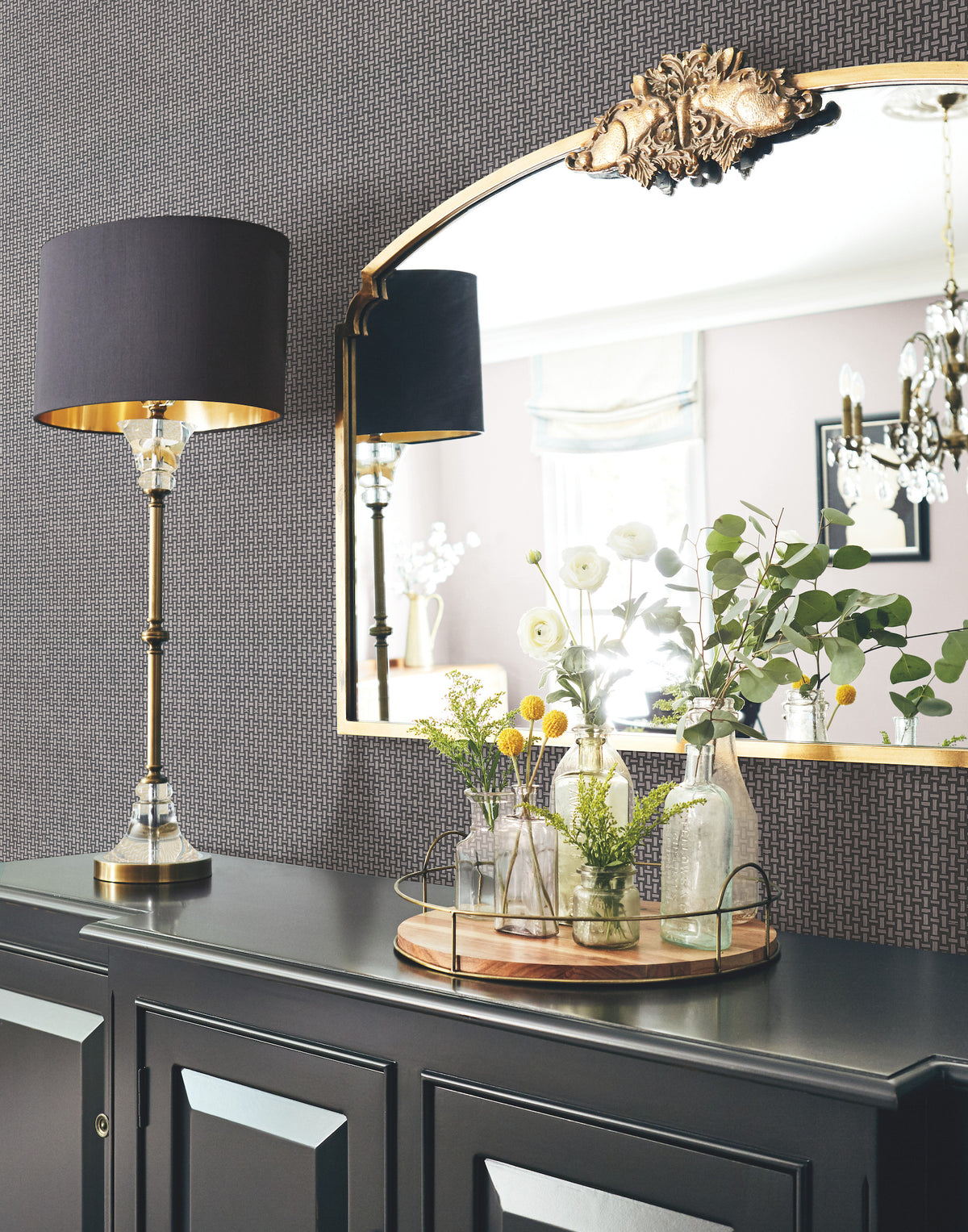 An elegant scene features Cottage Basket Wallpaper by York Wallcoverings as the backdrop, with a black sideboard beneath a gold-trimmed mirror. The arrangement is accented by a modern black lamp and a tray holding assorted glass vases filled with flowers and greenery, creating soft reflections in the mirror.