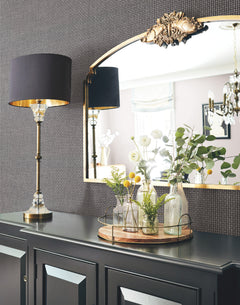 An elegant scene features Cottage Basket Wallpaper by York Wallcoverings as the backdrop, with a black sideboard beneath a gold-trimmed mirror. The arrangement is accented by a modern black lamp and a tray holding assorted glass vases filled with flowers and greenery, creating soft reflections in the mirror.