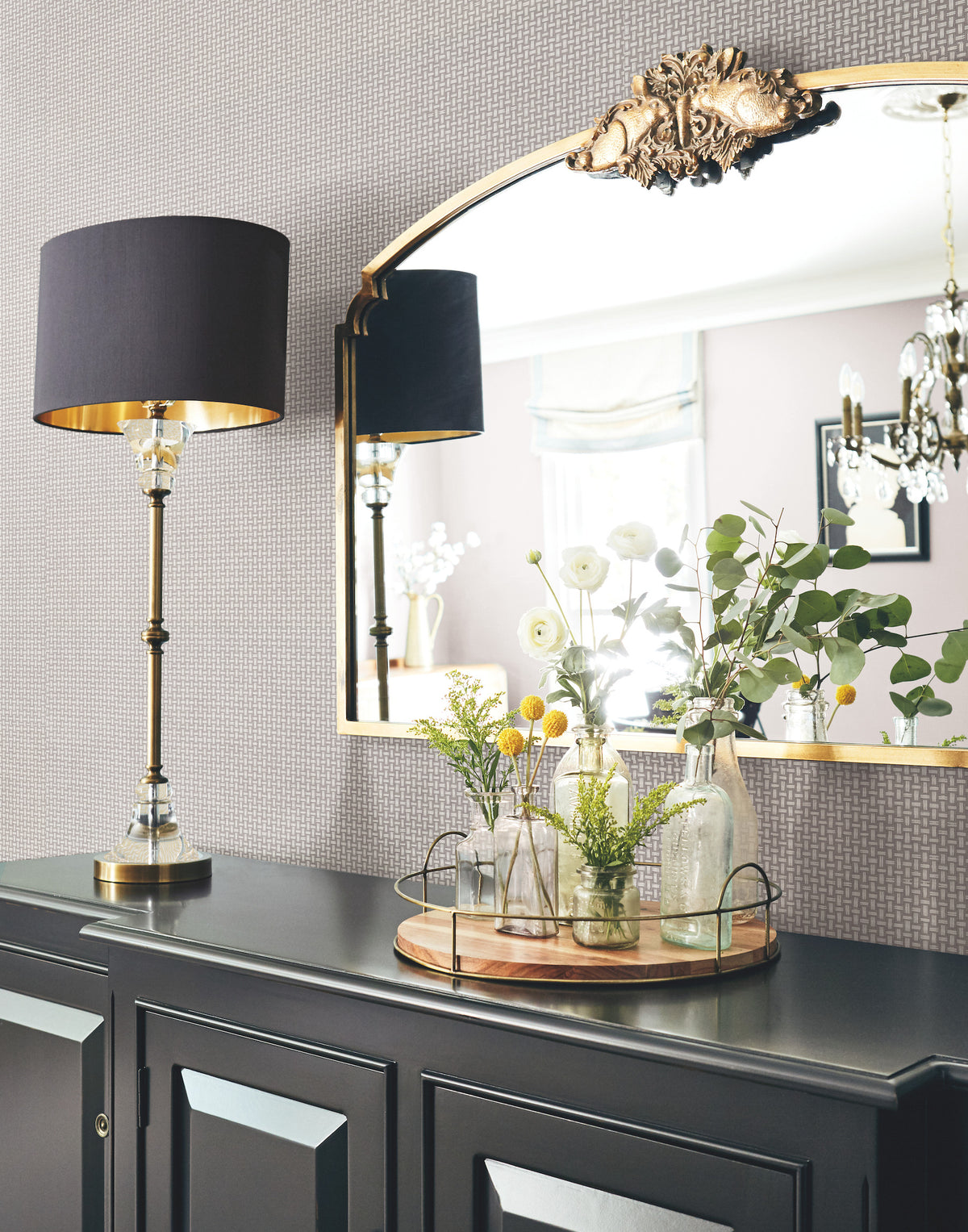 A black lamp, ornate gold mirror, and tray with glass vases holding flowers sit on a dark cabinet against a wall covered in Cottage Basket Wallpaper by York Wallcoverings.