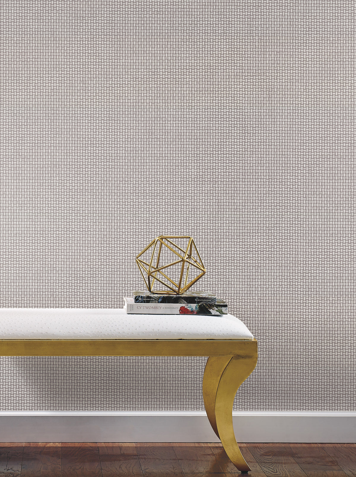A gold, geometric sculpture sits on two stacked books atop a white cushioned bench with gold legs, set against York Wallcoverings’ Cottage Basket Wallpaper and a dark wood floor.