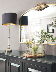 A black and gold table lamp sits on a dark cabinet beside a tray with glass vases of flowers. Above the cabinet, an ornate mirror reflects a chandelier and part of a window, all set against York Wallcoverings’ Cottage Basket Wallpaper.