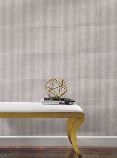 A white bench with gold legs sits on a wooden floor. On the bench are two books and a gold geometric sculpture. The background features Cottage Basket Wallpaper by York Wallcoverings, adding texture and a light tone to the space.