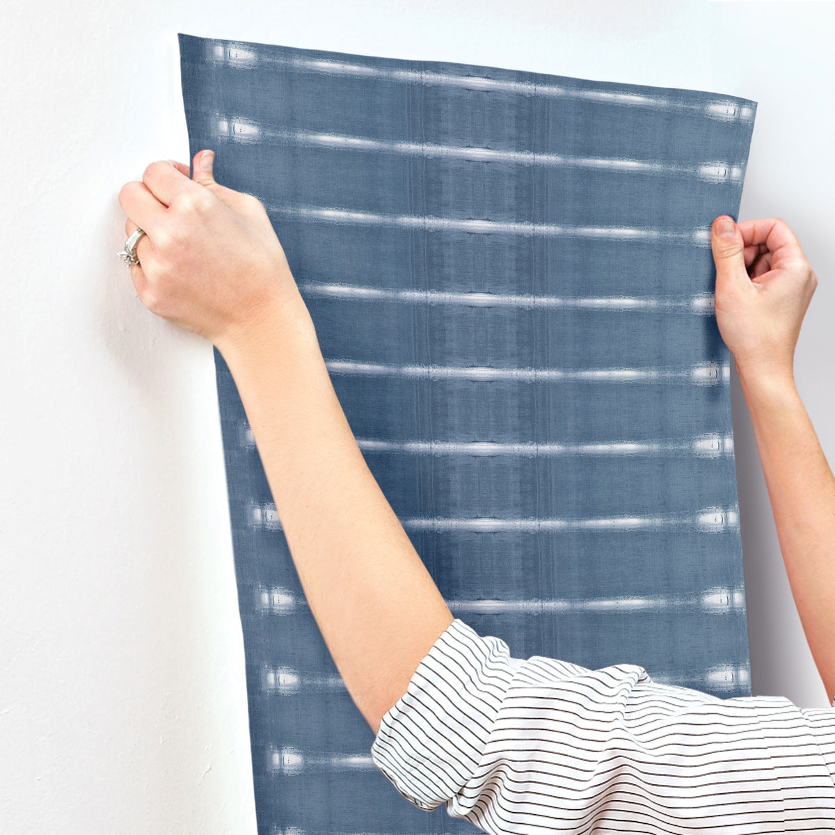 A person in a striped shirt is applying Resound Wallpaper by York Wallcoverings, featuring a blue plaid pattern, to a white wall and smoothing it out with both hands.
