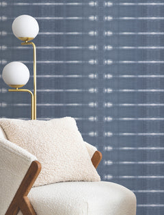 A cream armchair with a textured pillow sits next to a gold floor lamp with two globe lights, set against York Wallcoverings’ Resound Wallpaper, which features blue patterns with horizontal lines and dot accents.