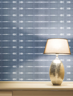 A metallic table lamp with a beige lampshade sits on a light-colored surface against a blue patterned wall featuring the Resound Wallpaper by York Wallcoverings.