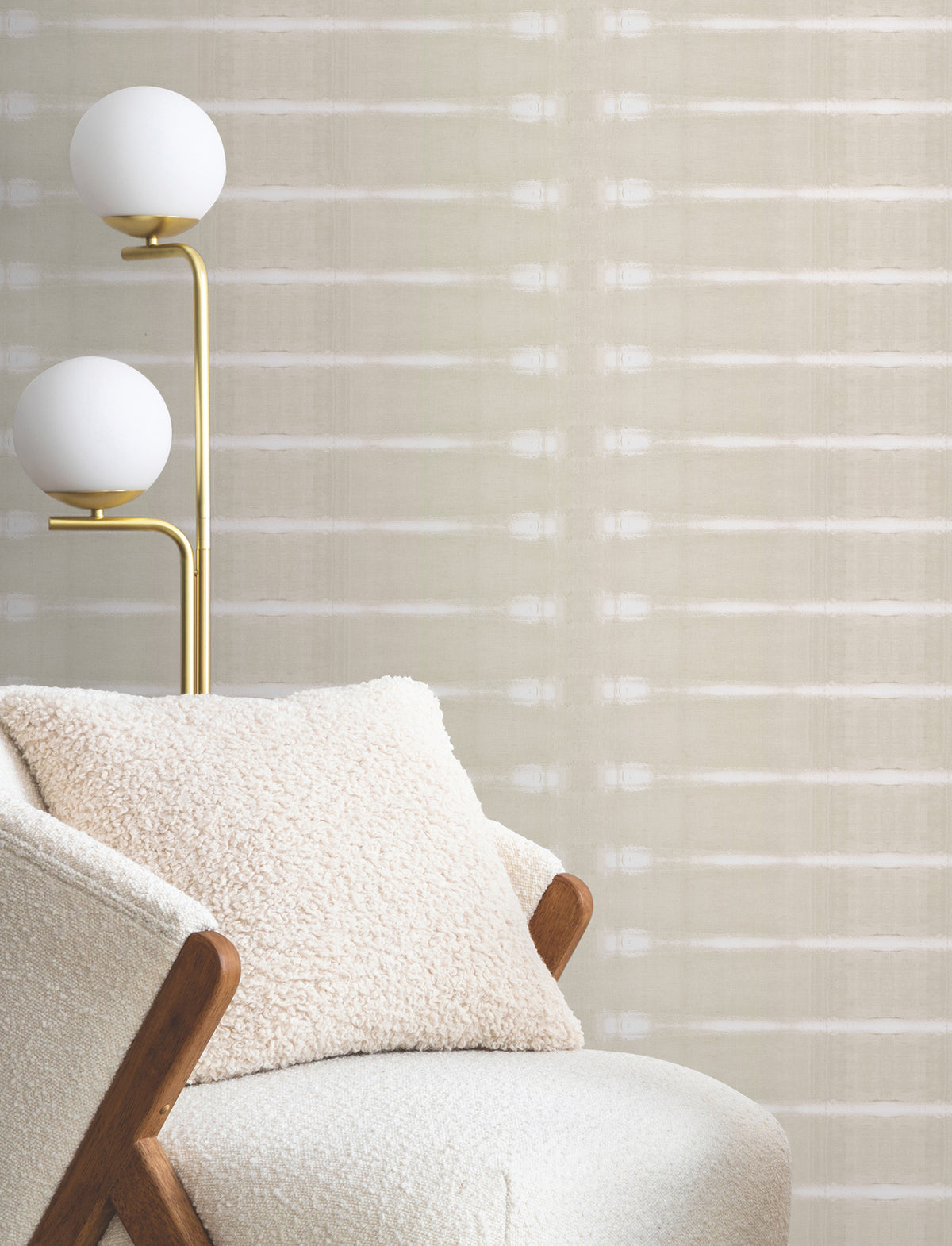 A cozy armchair with a textured pillow sits next to a modern brass floor lamp with two white globe lights, in front of a wall adorned with Resound Wallpaper by York Wallcoverings.