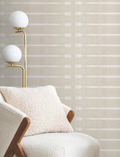 A cozy armchair with a textured pillow sits next to a modern brass floor lamp with two white globe lights, in front of a wall adorned with Resound Wallpaper by York Wallcoverings.