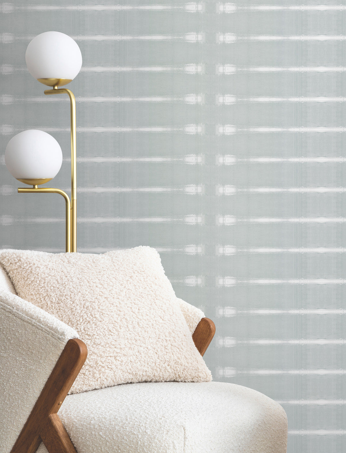 A cozy cream chair with a textured pillow sits beside a modern gold floor lamp with two globe lights, set against York Wallcoverings’ Resound Wallpaper in light gray.
