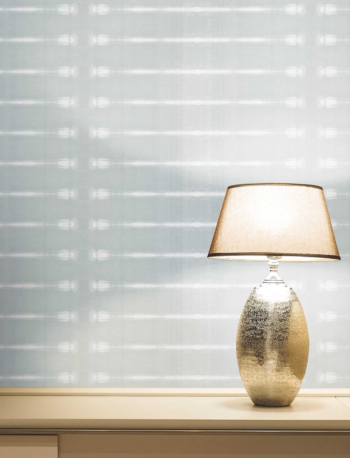 A lit table lamp with a metallic base sits on a light-colored surface against a pale Resound Wallpaper by York Wallcoverings, featuring subtle horizontal lines.