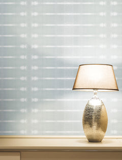 A lit table lamp with a metallic base sits on a light-colored surface against a pale Resound Wallpaper by York Wallcoverings, featuring subtle horizontal lines.