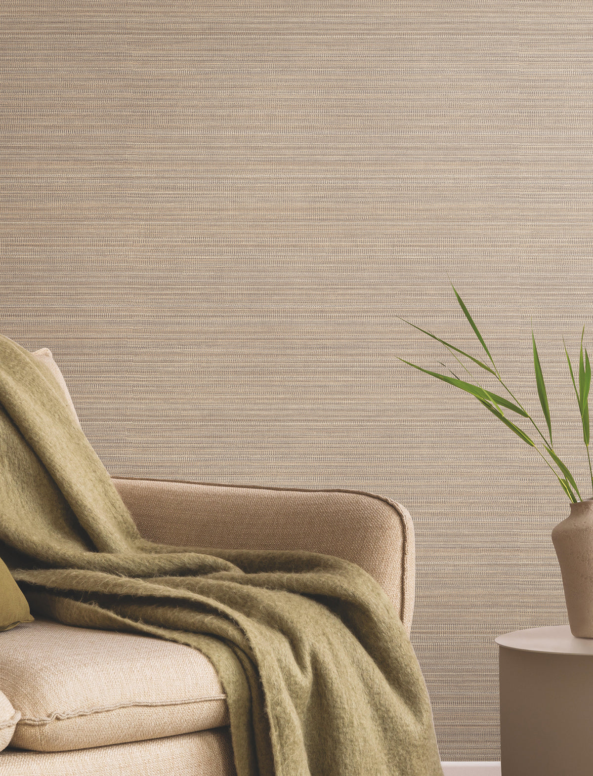 A beige armchair with a green throw blanket sits next to a small round table holding a simple vase with green leaves, set against a textured taupe wall featuring the Allineate Wallpaper by York Wallcoverings.