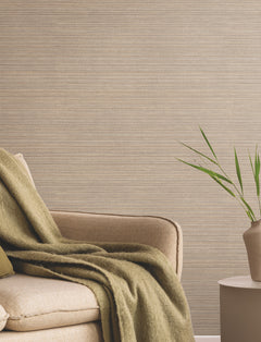 A beige armchair with a green throw blanket sits next to a small round table holding a simple vase with green leaves, set against a textured taupe wall featuring the Allineate Wallpaper by York Wallcoverings.