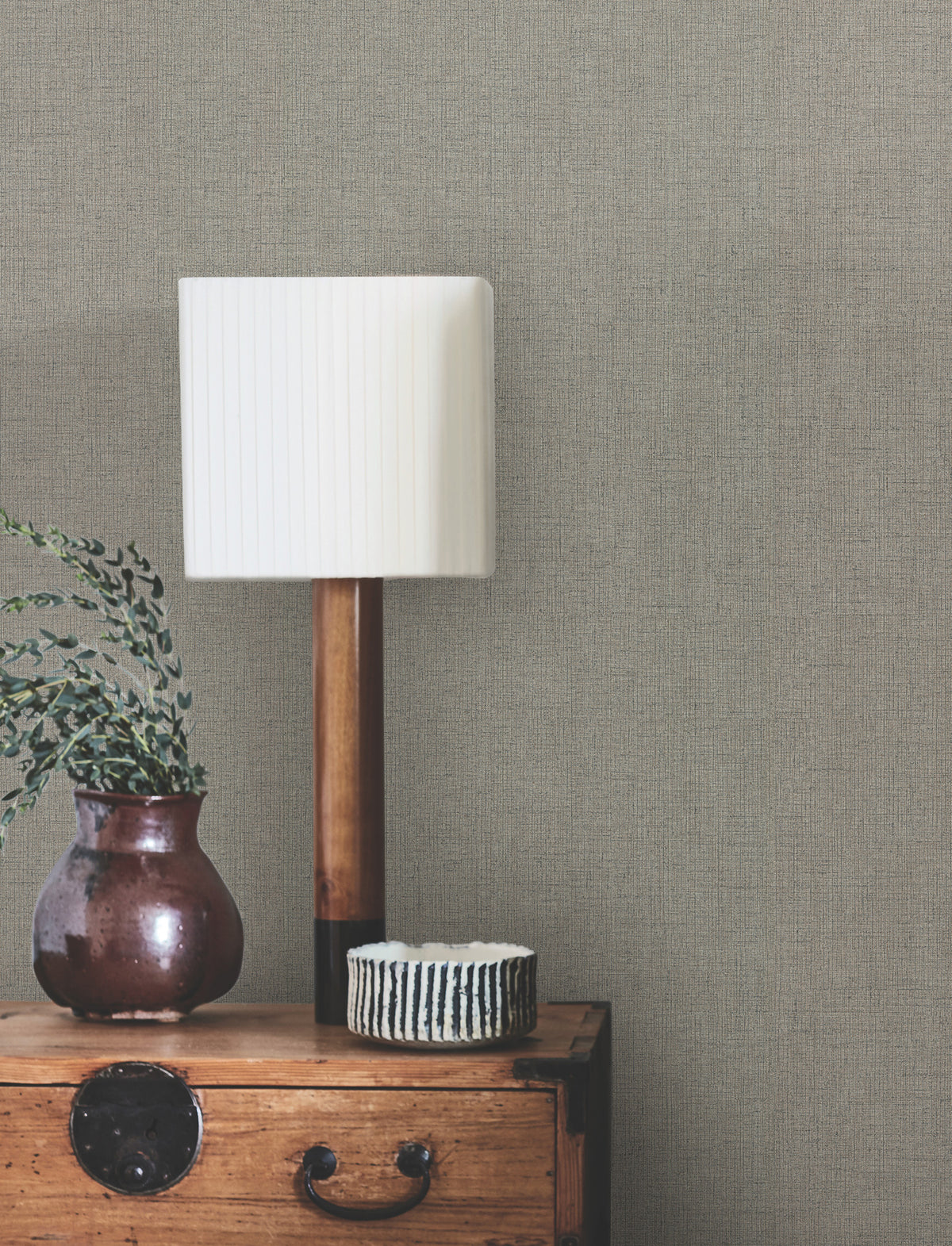 A white pleated lampshade on a wooden lamp, a brown vase with greenery, and a small striped bowl sit on a wooden chest against a wall covered with Hardy Linen Wallpaper by York Wallcoverings.