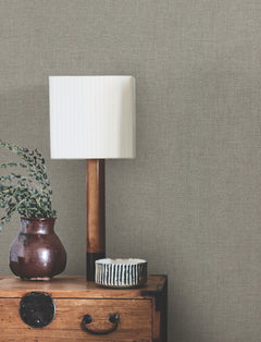 A white pleated lampshade on a wooden lamp, a brown vase with greenery, and a small striped bowl sit on a wooden chest against a wall covered with Hardy Linen Wallpaper by York Wallcoverings.