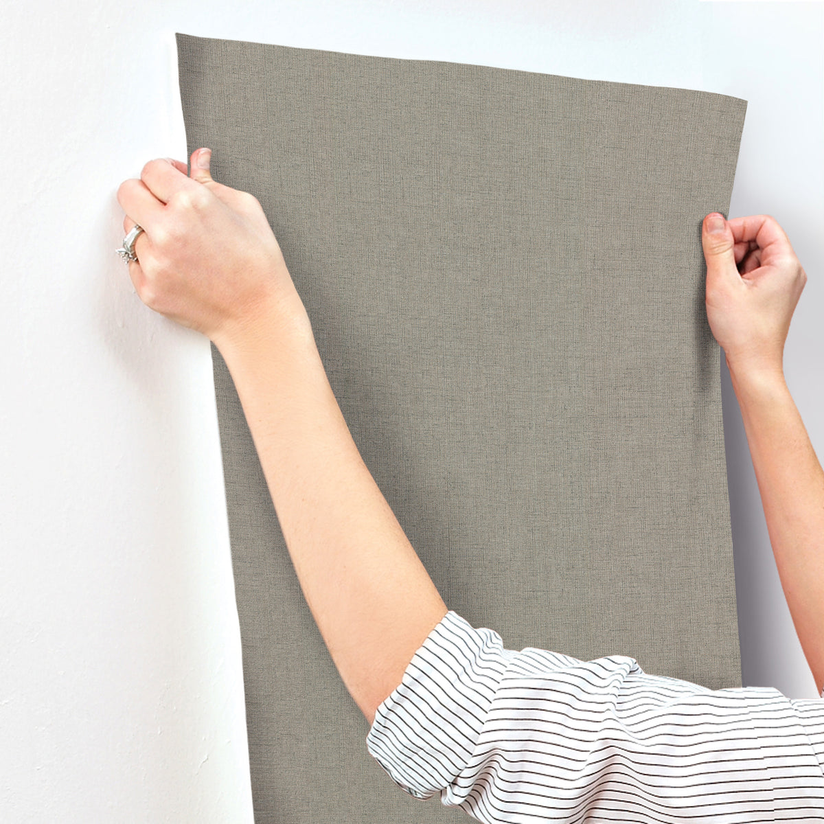 A person in a striped shirt uses both hands to hold up a piece of Hardy Linen Wallpaper by York Wallcoverings against a white wall.