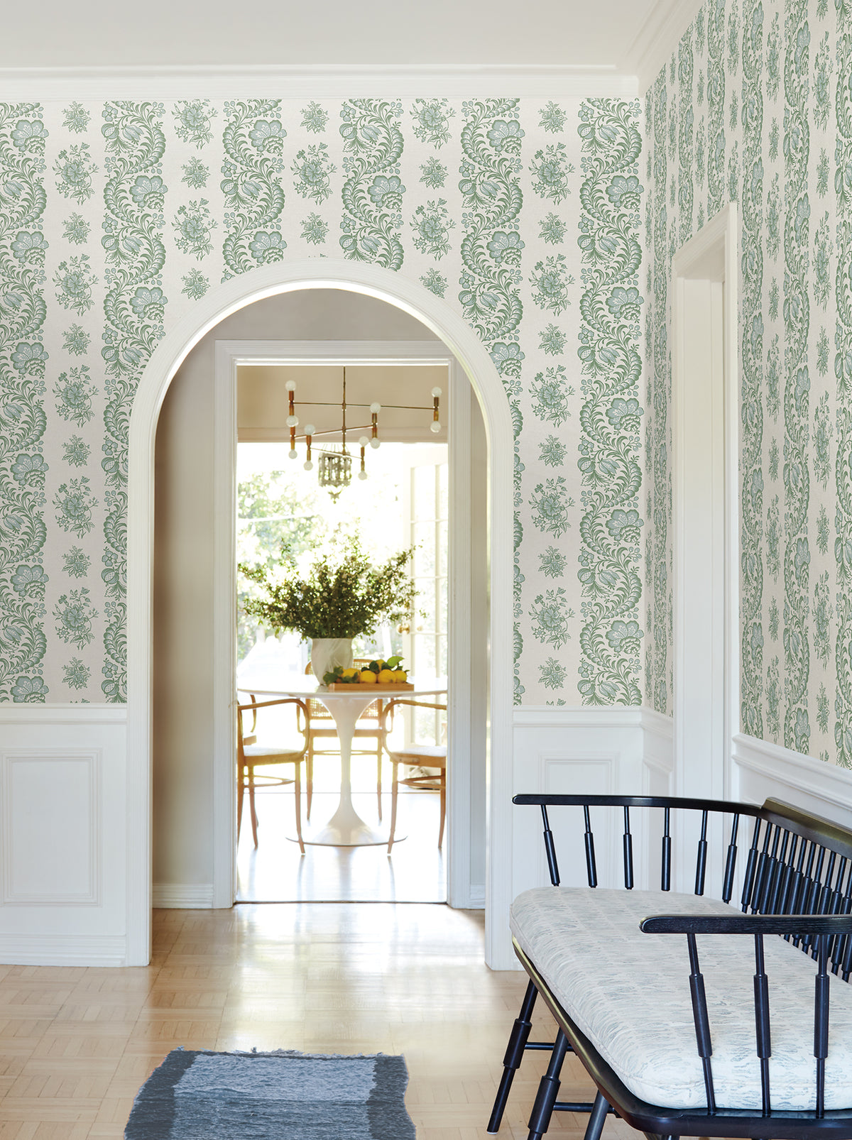A hallway adorned with Beaumont Floral Stripe Wallpaper by York Wallcoverings, featuring an arched doorway, a black bench with a light cushion, and a view into a bright dining room with a round table and chairs in the background.