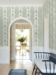 A hallway adorned with Beaumont Floral Stripe Wallpaper by York Wallcoverings, featuring an arched doorway, a black bench with a light cushion, and a view into a bright dining room with a round table and chairs in the background.