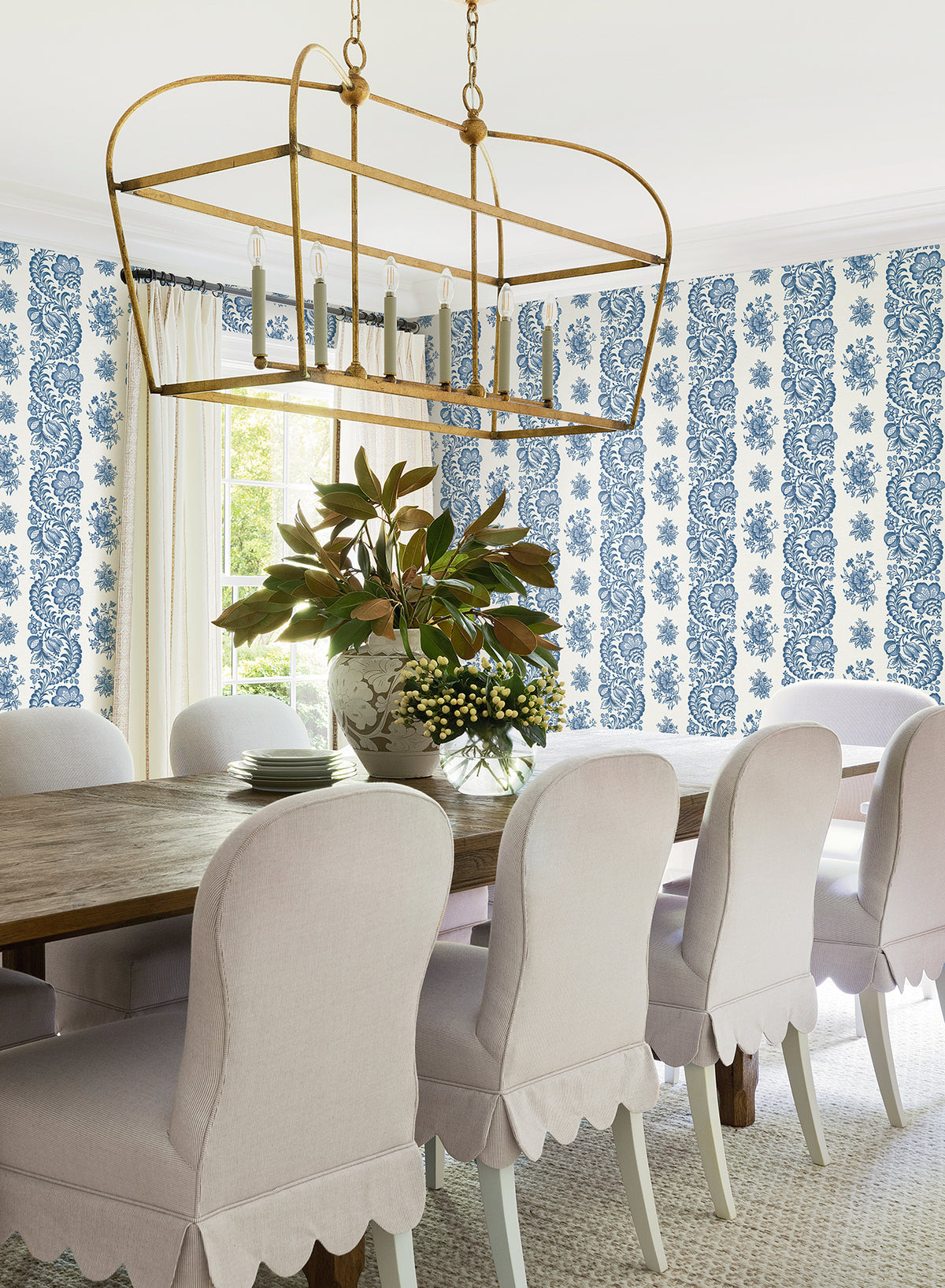 Bright dining room with a wooden table, eight white upholstered chairs, a large floral arrangement, gold chandelier, York Wallcoverings’ Beaumont Floral Stripe Wallpaper in blue and white, and natural light from a window.