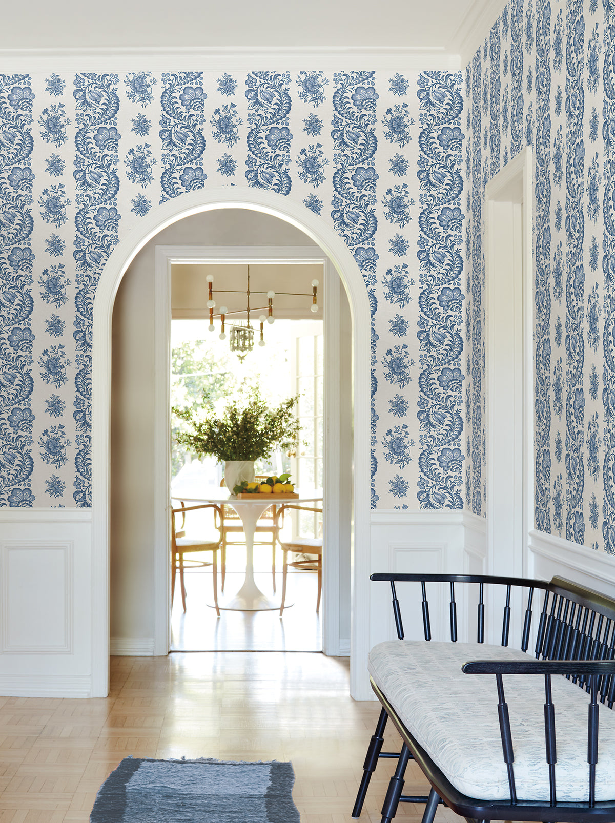 An arched doorway opens to a bright dining area with a white table and chairs. The hallway features Beaumont Floral Stripe Wallpaper by York Wallcoverings in blue and white, along with a black bench topped with a cushion and a blue rug on light wood floors.