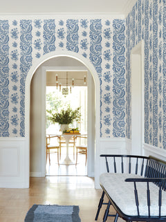 An arched doorway leads to a bright dining area with a round table and chairs. The walls are adorned with Beaumont Floral Stripe Wallpaper by York Wallcoverings, while a black bench with a cushion sits in the hallway.