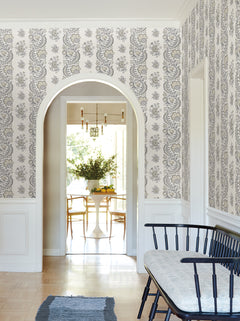 A bright hallway featuring York Wallcoverings’ Beaumont Floral Stripe Wallpaper, a black bench with a cushion, an arched doorway, and a view into a dining room with a round table, chairs, and a vase of flowers.