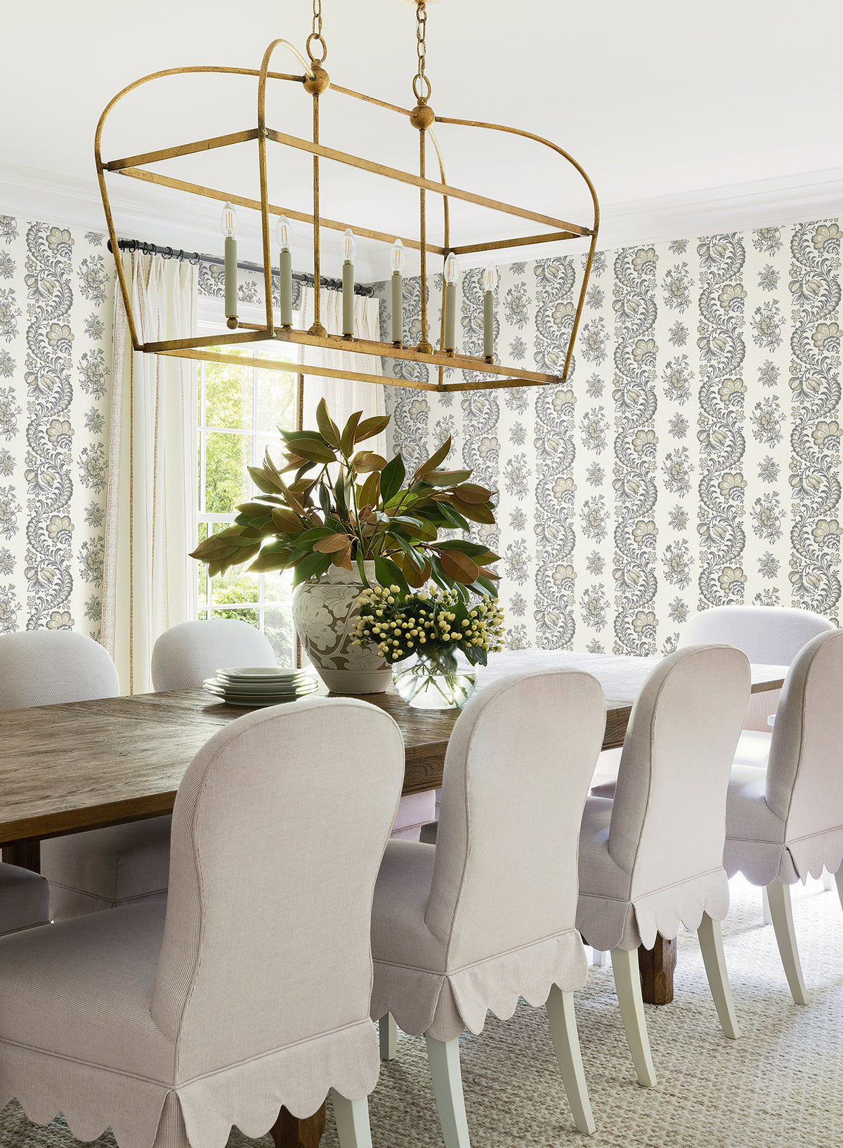 Sure! Here’s the revised sentence incorporating your product data: Elegant dining room with a wooden table, eight white scalloped chairs, a large gold chandelier, and a centerpiece of green leaves and white flowers. Sunlight streams in through a window, highlighting the Beaumont Floral Stripe Wallpaper by York Wallcoverings.