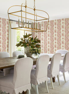 A dining room featuring a wooden table, nine white upholstered chairs, Beaumont Floral Stripe Wallpaper by York Wallcoverings, a large gold chandelier, and two vases of flowers as centerpieces. Sunlight streams in through a window with white curtains.