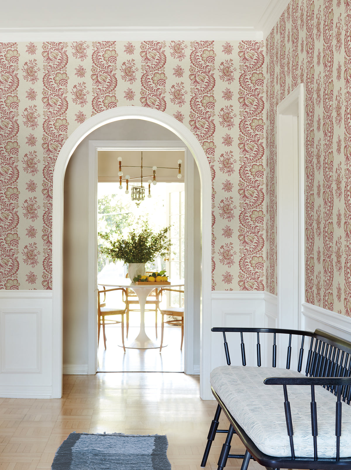 A hallway adorned with York Wallcoverings’ Beaumont Floral Stripe Wallpaper features an arched doorway, a black bench with a cushion, and offers a view into a dining room with a round table, chairs, and a vase of flowers.