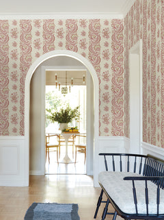 A hallway adorned with York Wallcoverings’ Beaumont Floral Stripe Wallpaper, featuring an arched doorway that opens to a dining area with a white table and chairs, and a black bench with a cushion set beside a white wainscoted wall, all illuminated by natural light.