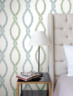A bedside table with books, eyeglasses, and a lamp sits next to a tufted beige headboard. The wall features the Laureate Vine Trellis Wallpaper by York Wallcoverings in elegant green and blue tones.