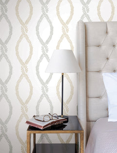 A bedside table with books and glasses, a lamp with a white shade, sits beside a tufted beige headboard and white pillow, with Laureate Vine Trellis Wallpaper by York Wallcoverings in the background.