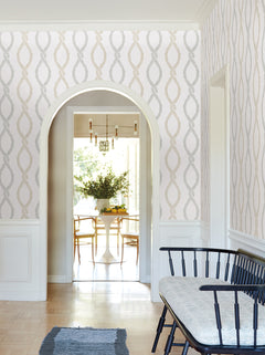 A hallway adorned with Laureate Vine Trellis Wallpaper by York Wallcoverings features an arched doorway, a black bench with cushion, and offers a view into a dining area with a round table, chairs, and a vase of green foliage. Sunlight streams in from the windows in the background.
