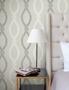 A bedside table with a lamp, books, and eyeglasses sits next to a beige tufted headboard and York Wallcoverings’ Laureate Vine Trellis Wallpaper.