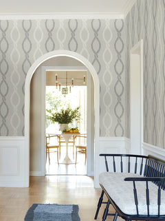 A light-filled dining room is seen through an arched doorway, featuring York Wallcoverings’ Laureate Vine Trellis Wallpaper, a black bench with a cushion, a blue rug, and a white oval table with chairs and a vase of flowers.