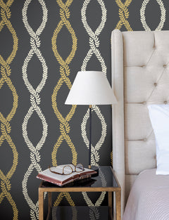 A beige bed with a tufted headboard sits beside a glass-top nightstand displaying books, glasses, and a lamp. The wall features York Wallcoverings’ Laureate Vine Trellis Wallpaper, showcasing an elegant gold and cream leaf pattern on a dark background.