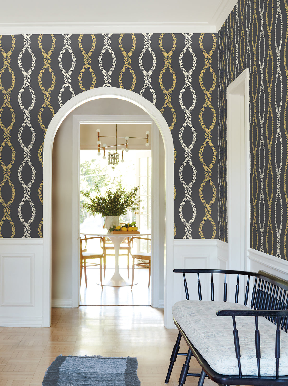 A hallway adorned with York Wallcoverings’ Laureate Vine Trellis Wallpaper, featuring a white archway, a wooden bench with cushion, and a view into a bright dining room with a round table and chairs near a large window.